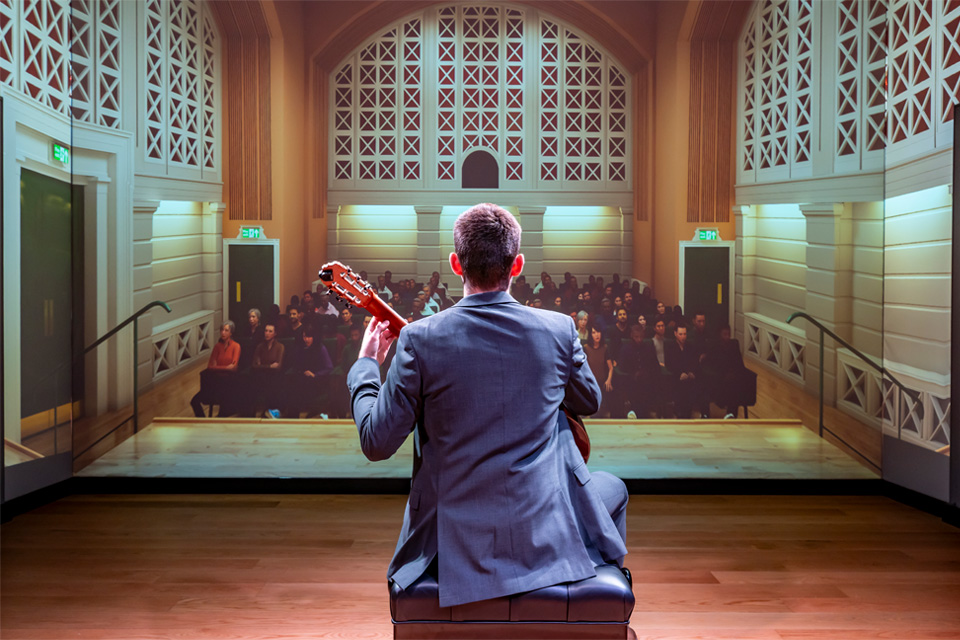 A male student wearing a suit, performing on a guitar in front of a fake audience in a visual simulator in front of him.